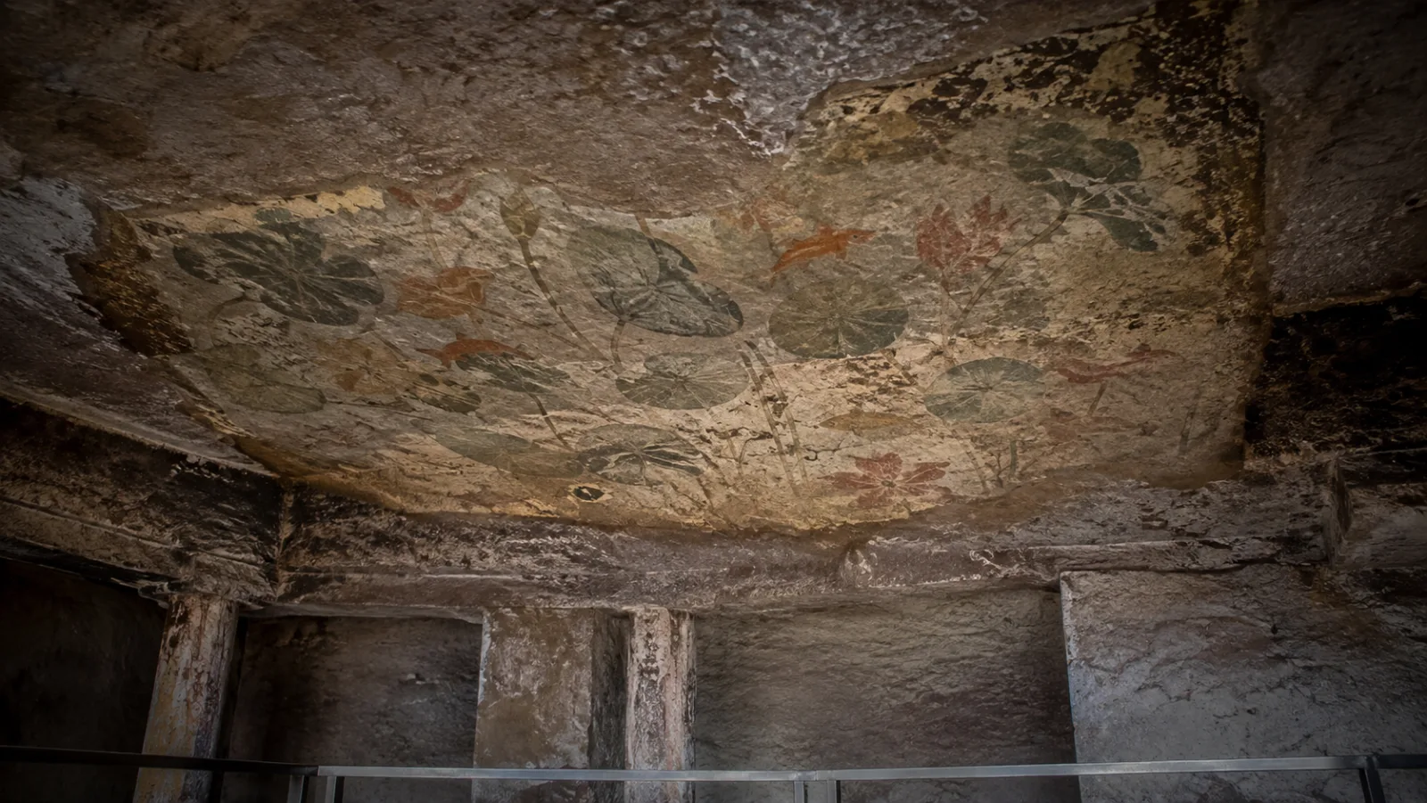 Editorial interpretation of a fragile lotus-pool ceiling mural above weathered rock-cut pillars and a conservation barrier