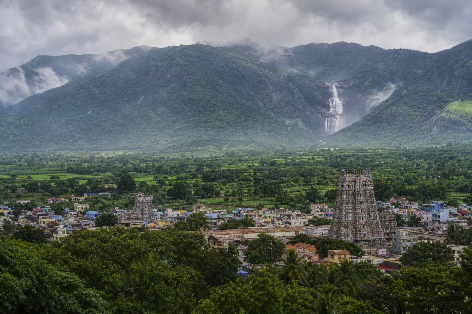 Editorial landscape interpretation of a Tenkasi temple town, green Western Ghats and a distant seasonal waterfall